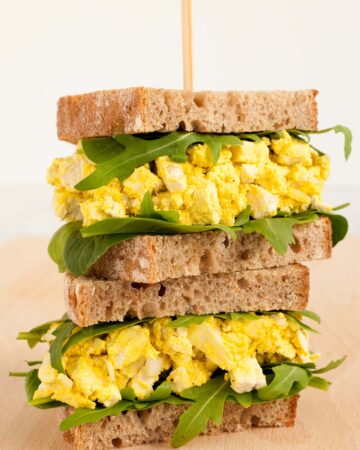 A stack of two sandwiches. Both with brown sliced bread, arugula, and tofu cubes in thick yellow sauce to mimic egg salad.