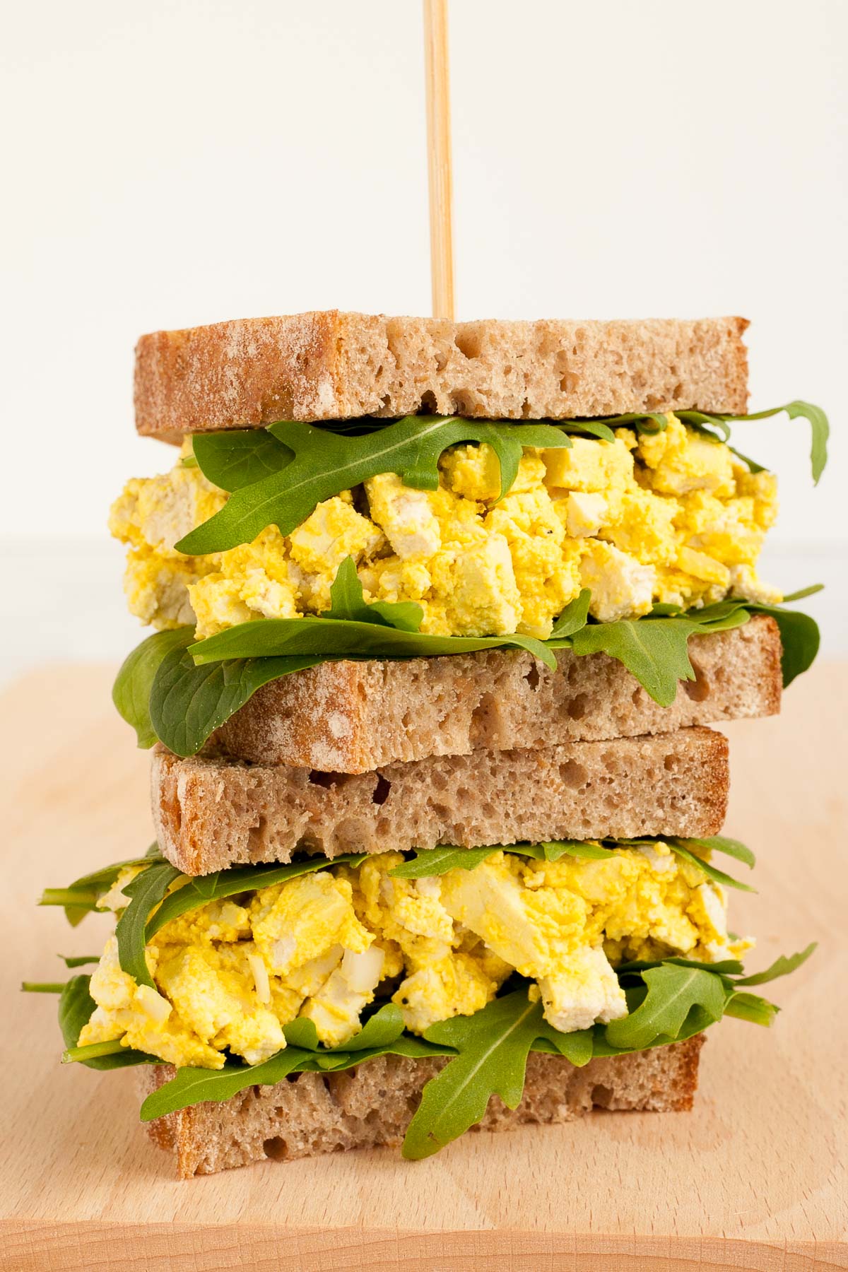 A stack of two sandwiches. Both with brown sliced bread, arugula, and tofu cubes in thick yellow sauce to mimic egg salad.