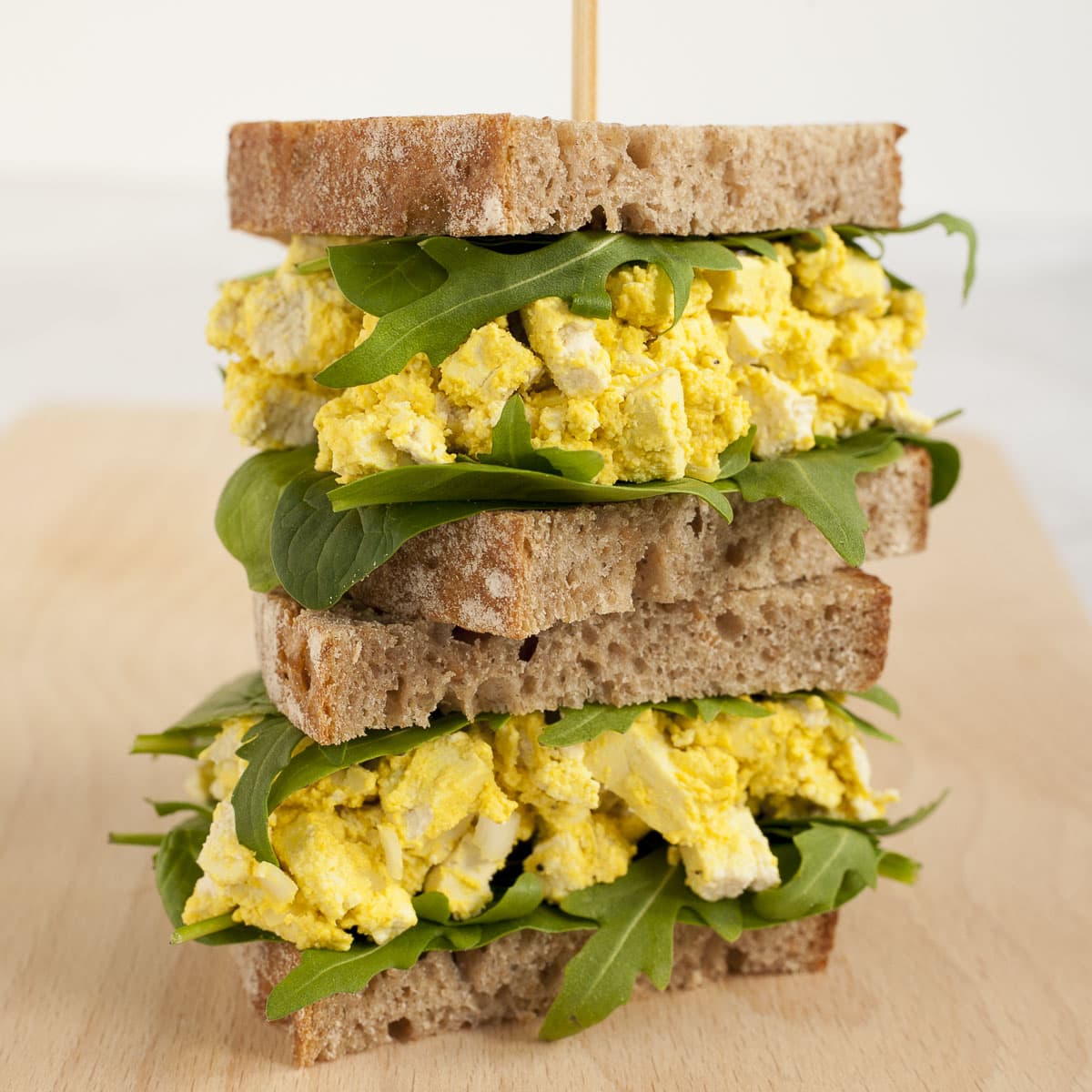 A stack of two sandwiches. Both with brown sliced bread, arugula, and tofu cubes in thick yellow sauce to mimic egg salad.