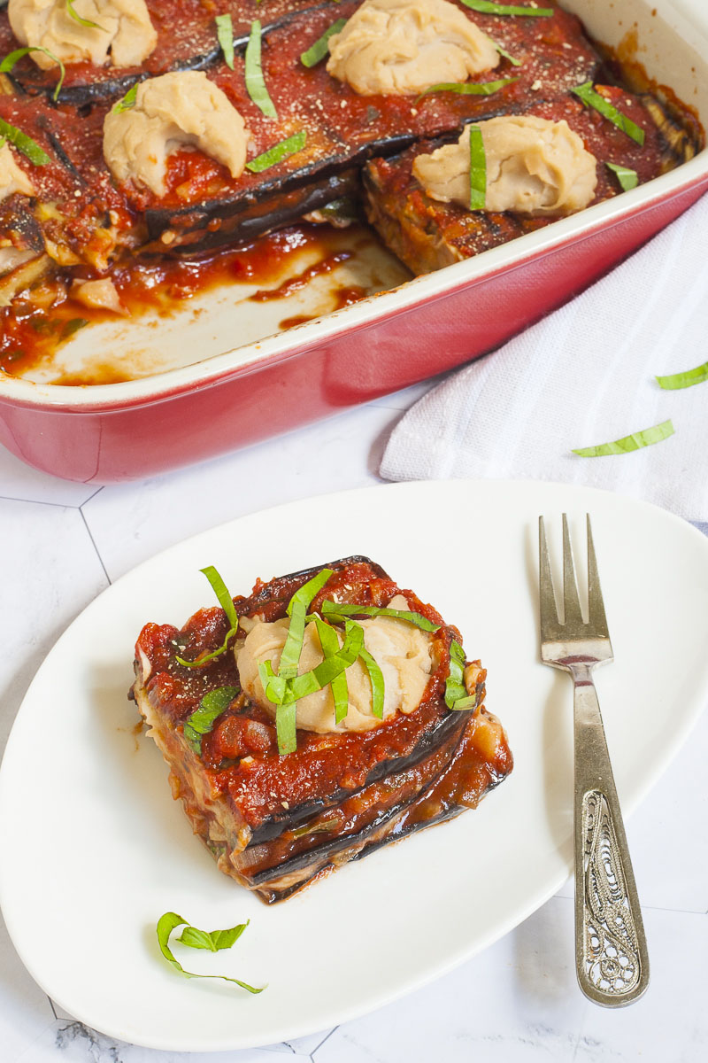 3 slices of purple eggplant in between red and white sauces on a white plate topped with fresh green herbs. In the background there is a red casserole with the remaining eggplant parmesan