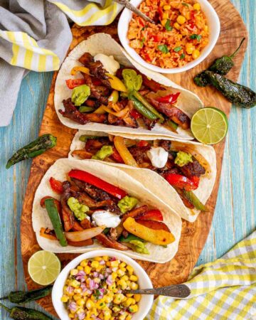 Small tortillas with colorful pepper slices and crispy brown mushroom shreds topped with white and green sauces on a wooden board. Surrounded by side dishes.
