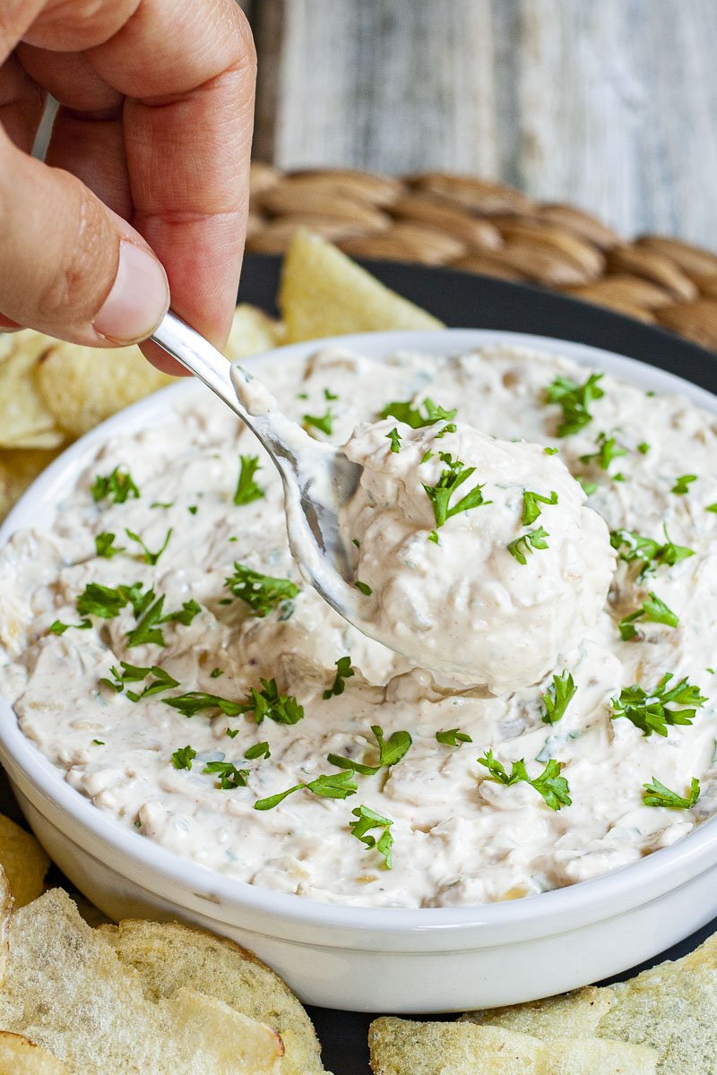 White bowl with white thick creamy sauce in it sprinkled with fresh green herbs. A hand is holding a spoon and taking a bit from the middle.