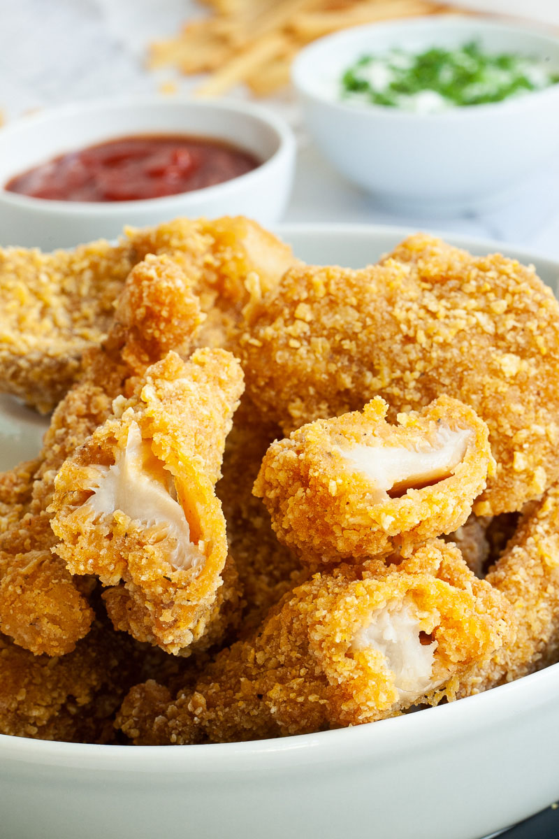 Brown breaded nuggets in a round plate. A couple of pieces are cut in the middle to see the white flesh in the middle. French fries to the side with two small bowls. One with ketchup and the other is a white sauce with chopped green herbs.