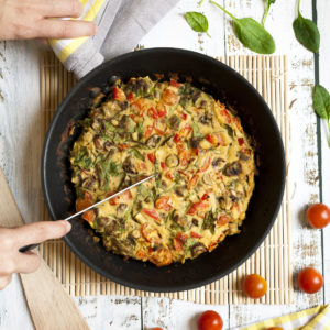 A frying pan with frittata, chopped veggies in an eggy batter. A hand is holding a knife to make slices.
