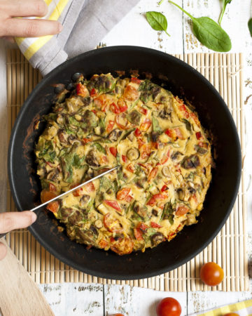 A frying pan with frittata, chopped veggies in an eggy batter. A hand is holding a knife to make slices.