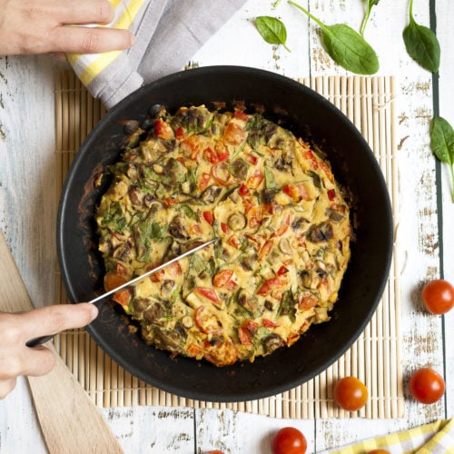 A frying pan with frittata, chopped veggies in an eggy batter. A hand is holding a knife to make slices.
