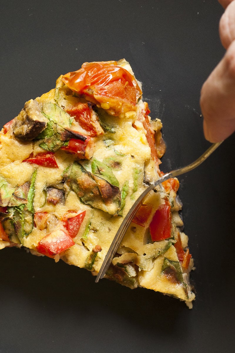 A black plate with a slice of yellow frittata full of veggies like chopped red bell pepper, green spinach leaves, brown mushroom, red tomatoes. A hand is holding a fork and about to take a bite.