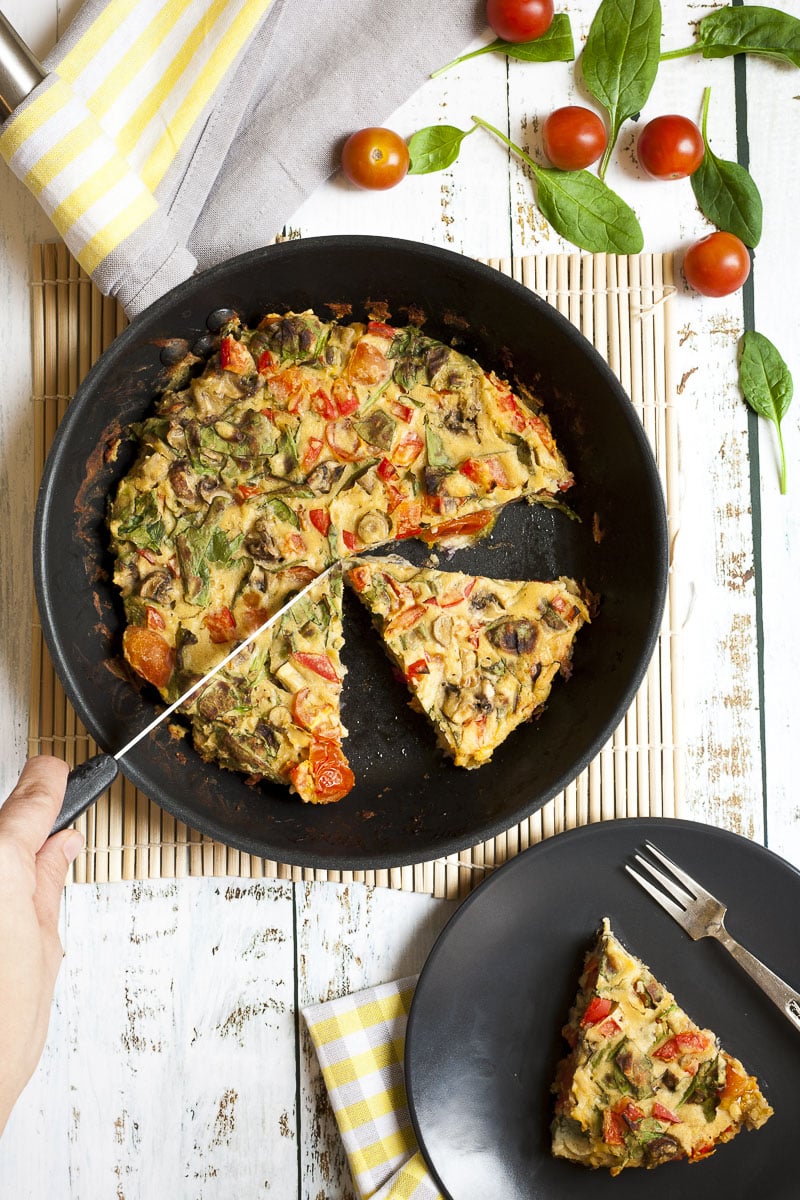Frying pan from above with a yellow frittata full of veggies like chopped red bell pepper, green spinach leaves, brown mushroom, red tomatoes. A hand is holding a knife to make another slice.