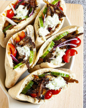 Several pita pocket on a white serving tray stuffed with crispy brown shredded oyster mushroom, tomato and cucumber slices andwhite-green tzaztziki sauce