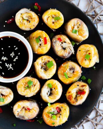 Lots of round-shaped rice paper dumplings arranged in a semi-circular dish facing upwards sprinkled with sesame seeds, chopped spring onion and red hot pepper around a brown dipping sauce.