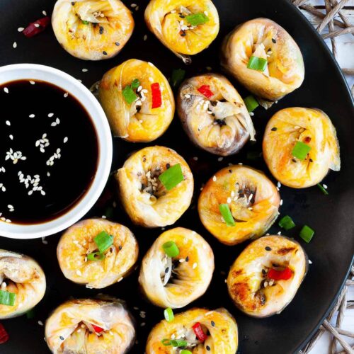 Lots of round-shaped rice paper dumplings arranged in a semi-circular dish facing upwards sprinkled with sesame seeds, chopped spring onion and red hot pepper around a brown dipping sauce.