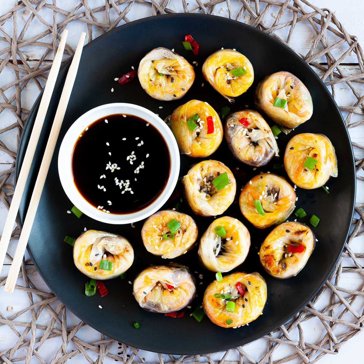 Lots of round-shaped rice paper dumplings arranged in a semi-circular dish facing upwards sprinkled with sesame seeds, chopped spring onion and red hot pepper around a brown dipping sauce. Wooden chopsticks are on the side.