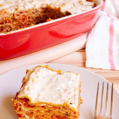 A slice of layered lasagna is served on a plate. A red casserole dish with the remaining lasagna is right behind it.
