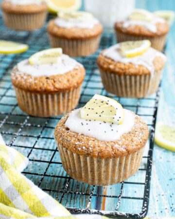 Several lemon poppy seed muffins on a black wire rack topped with a white glaze and a small lemon slices, and sprinkled with poppy seeds. Lemon slices are scattered around them.