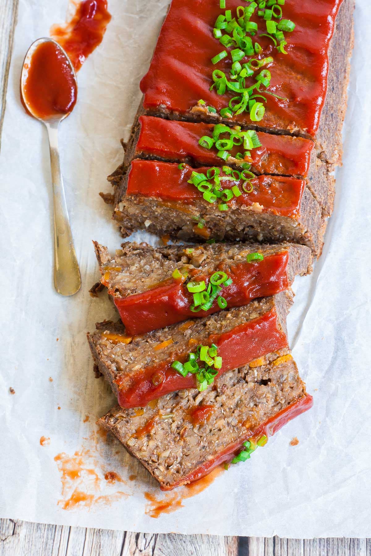 A long brown lentil loaf on parchment paper has been cut into slices. 3 slices has fallen in the front. All slices are glazed with red sauce and topped with chopped scallions.