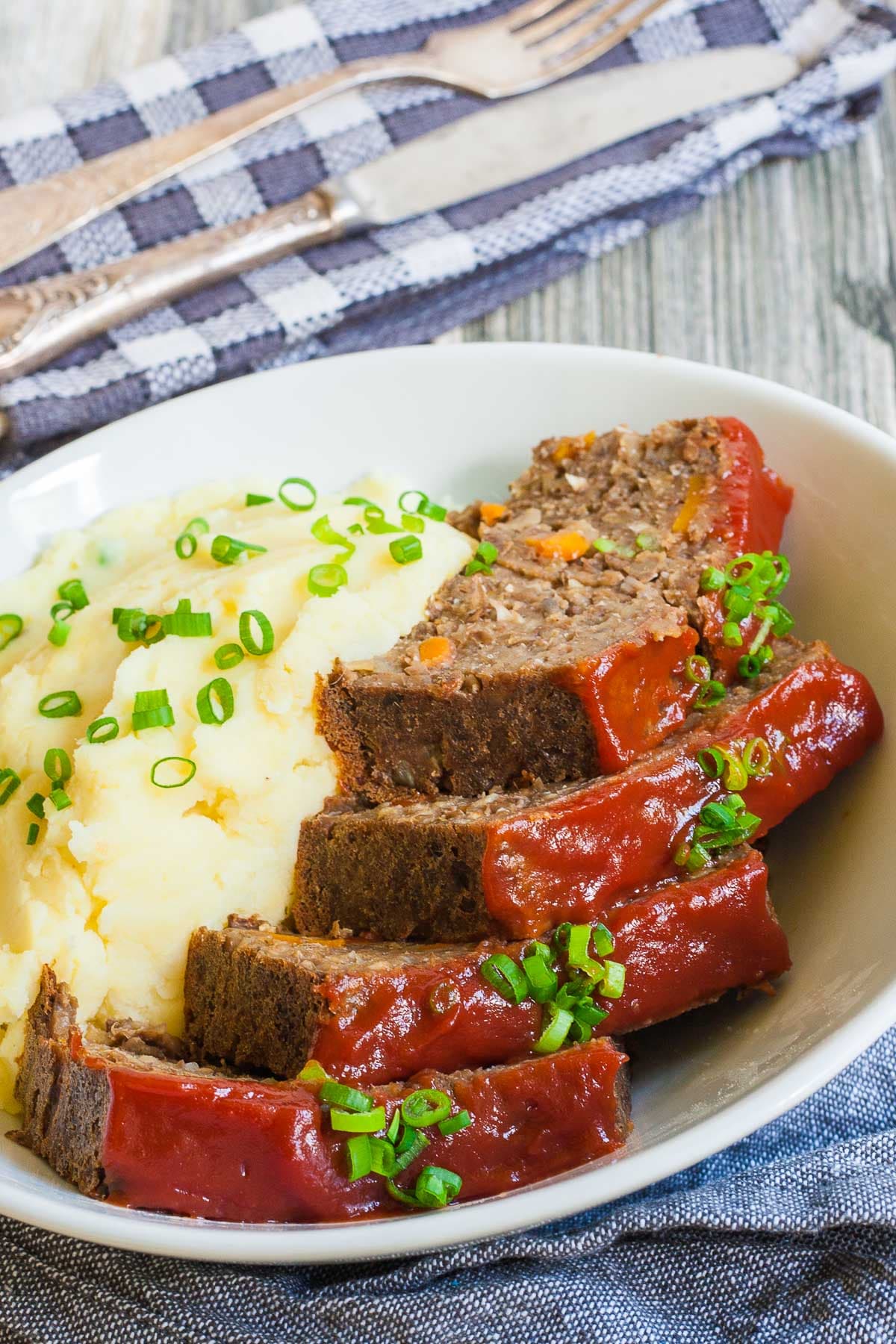 The Perfect Vegan Lentil Loaf should be deliciously rich, moist and juicy, and easy to make. Well, we give you a 10-ingredient healthy, hearty, rich and flavorful vegan lentil loaf that is not only gluten-free, but grain-free as well. #veganlentilloaf #lentiloaf #vegetarianlentilloaf #grainfree #glutenfree 4 slices of lentil loaf glazed with a red sauce served in a large bowl with mashed potatoes and chopped scallions.