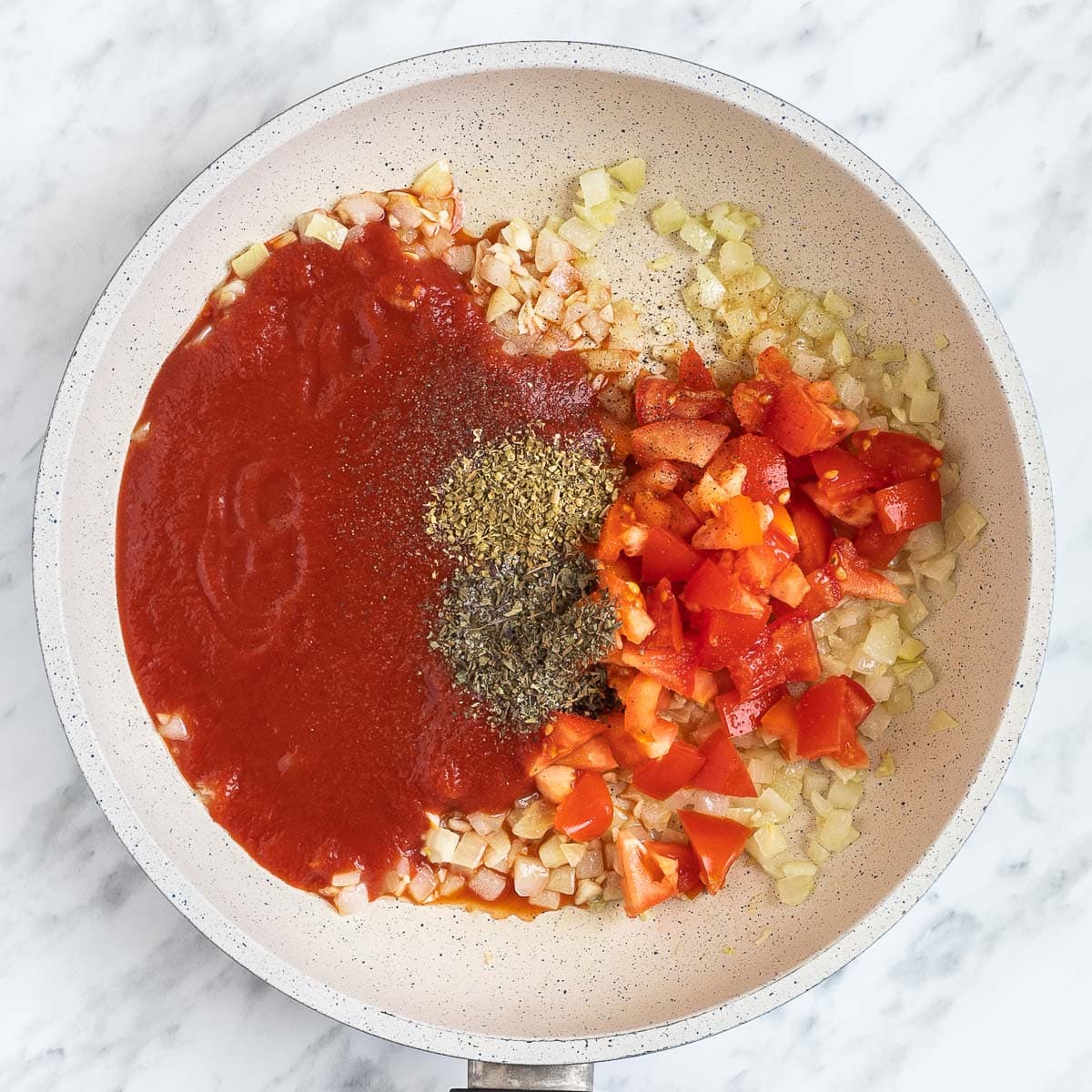 White frying pan with chopped onion, garlic, tomato sauce, chopped tomatoes and dried herbs.