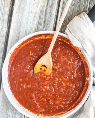 A white frying pan with red chunky sauce. A smiling wooden spatula is placed inside.