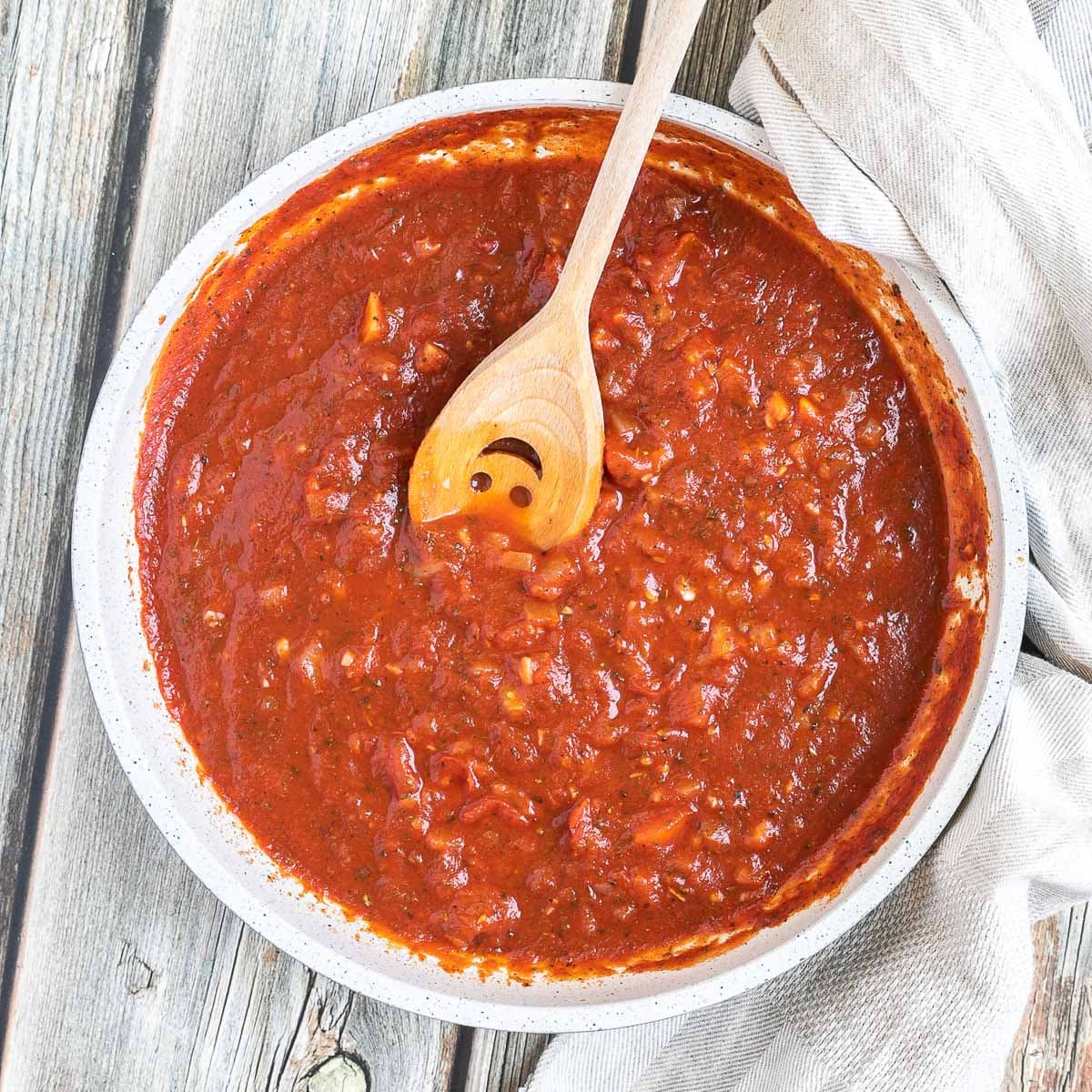 A white frying pan with red chunky sauce. A smiling wooden spatula is placed inside.
