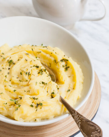 Mashed potatoes sprinkled with green herbs in a white bowl with a spoon. A small gravy boat is right behind it.