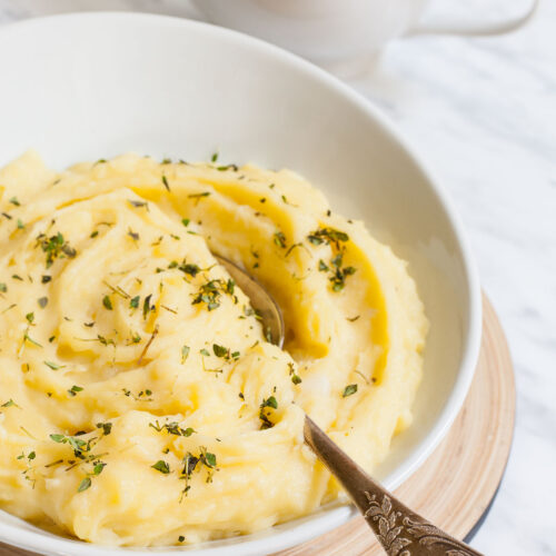 Mashed potatoes sprinkled with green herbs in a white bowl with a spoon. A small gravy boat is right behind it.