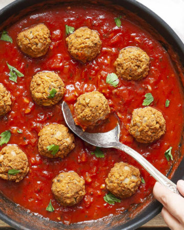 A skillet from above with several baked brown meatballs in bright red sauce and chopped green herbs. A spoon is taking one ball from the middle.