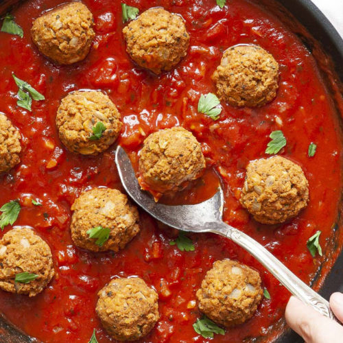 A skillet from above with several baked brown meatballs in bright red sauce and chopped green herbs. A spoon is taking one ball from the middle.