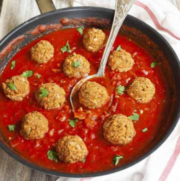 A skillet from above with several baked brown meatballs in bright red sauce and chopped green herbs. A spoon is taking one ball from the middle.