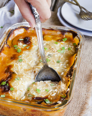 The corner of a glass dish with a serving spoon. The dish is full of yellow-red-brown food topped with chopped parsley.