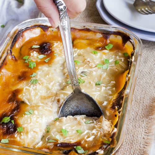 The corner of a glass dish with a serving spoon. The dish is full of yellow-red-brown food topped with chopped parsley.