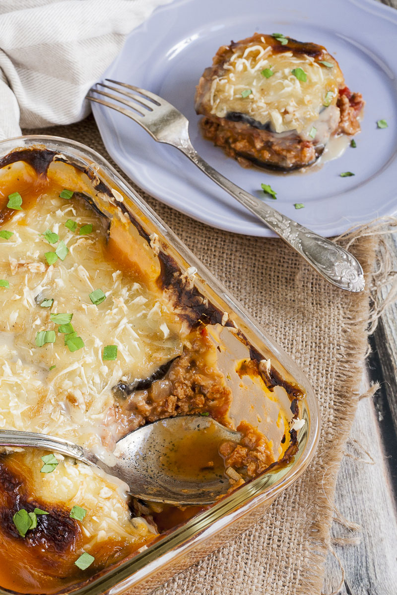 A glass dish with a serving spoon. The dish is full of yellow-red-brown food topped with chopped green herbs. One serving of vegan moussaka is on a light purple plate.