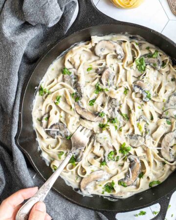 Cast iron skillet with tagliatelle in a creamy light brown sauce with sliced mushrooms and freshly chopped green herbs. A hand is holding a fork to the middle.