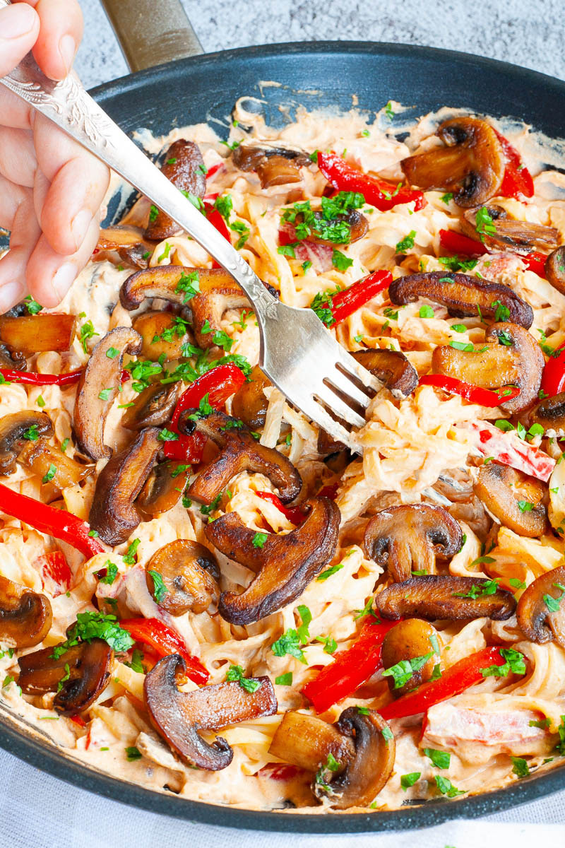 Frying pan from above with fettuccini pasta in a white-orange creamy sauce with sliced crispy mushrooms, red bell pepper sticks and freshly chopped parsley.