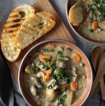 Grayish brown stew with mushroom slices, carrots and potatoes served in a brown bowl. 2 slice of crusty bread is placed next to it.