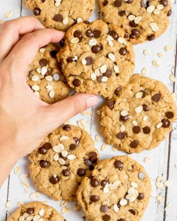 Lots of cookies with oats and chocolate chips on a wooden surface. More oats are sprinkled around them. A hand is taking one cookie for the middle.
