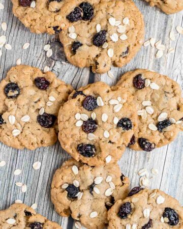 Lots of cookies with oats and raisins on a wooden surface. More oats are sprinkled around them.