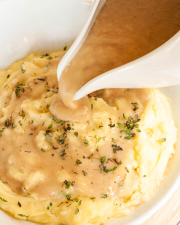 White bowl with mashed potatoes. Light brown gravy is pouring on top of it from a gravy boat.