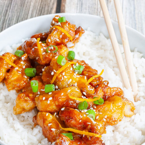 A large white bowl with white rice and sticky brown battered strips on top sprinkled with sesame seeds, orange peels and chopped spring onion