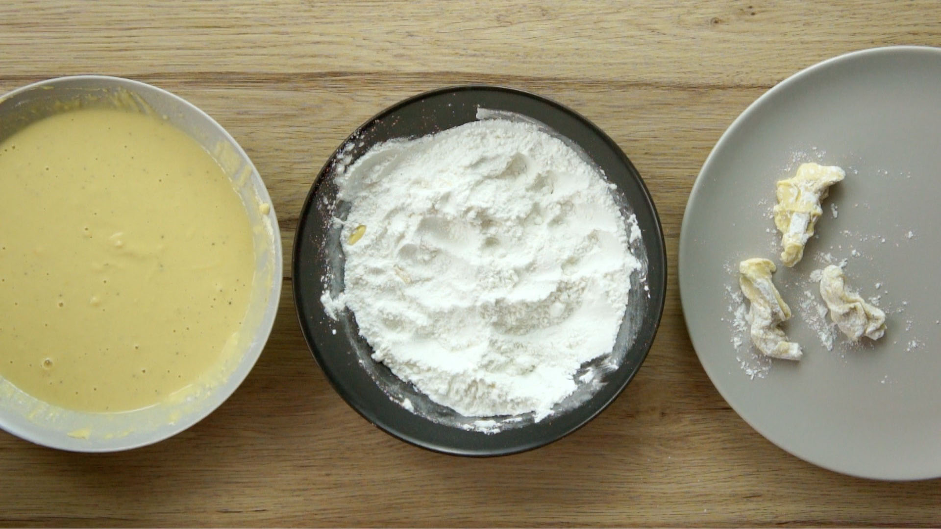 3 bowls from above. The first has a yellow batter, the second has white flour and the thirds with 3 battered mushroom strips.