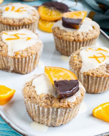 Several orange muffins on a light blue plate topped with a white glaze and orange slices partly dipped in chocolate or white glaze with orange zest pieces. Orange slices are scattered around them.
