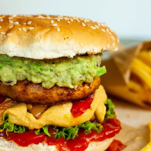 A burger with ketchup, lettuce, yellow cheese sauce, bell peppers, guacamole and grilled mushroom cap on brown paper. French fries are placed next to it.