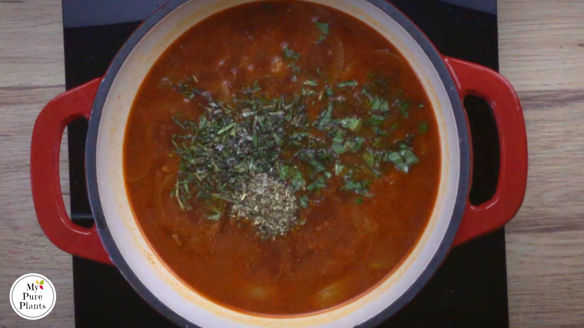 Red-white enameled Dutch oven with a thin red soup and heaps of dried and freshly chopped herbs in the middle.