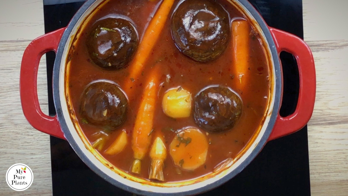 Red-white enameled Dutch oven with a thin red soup and 4 large portobello mushroom caps, whole baby carrots, and half potatoes.