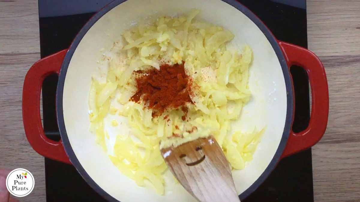 Red-white enameled Dutch oven with lightly cooked sliced onion and garlic, and a small heap of red powder in the middle.