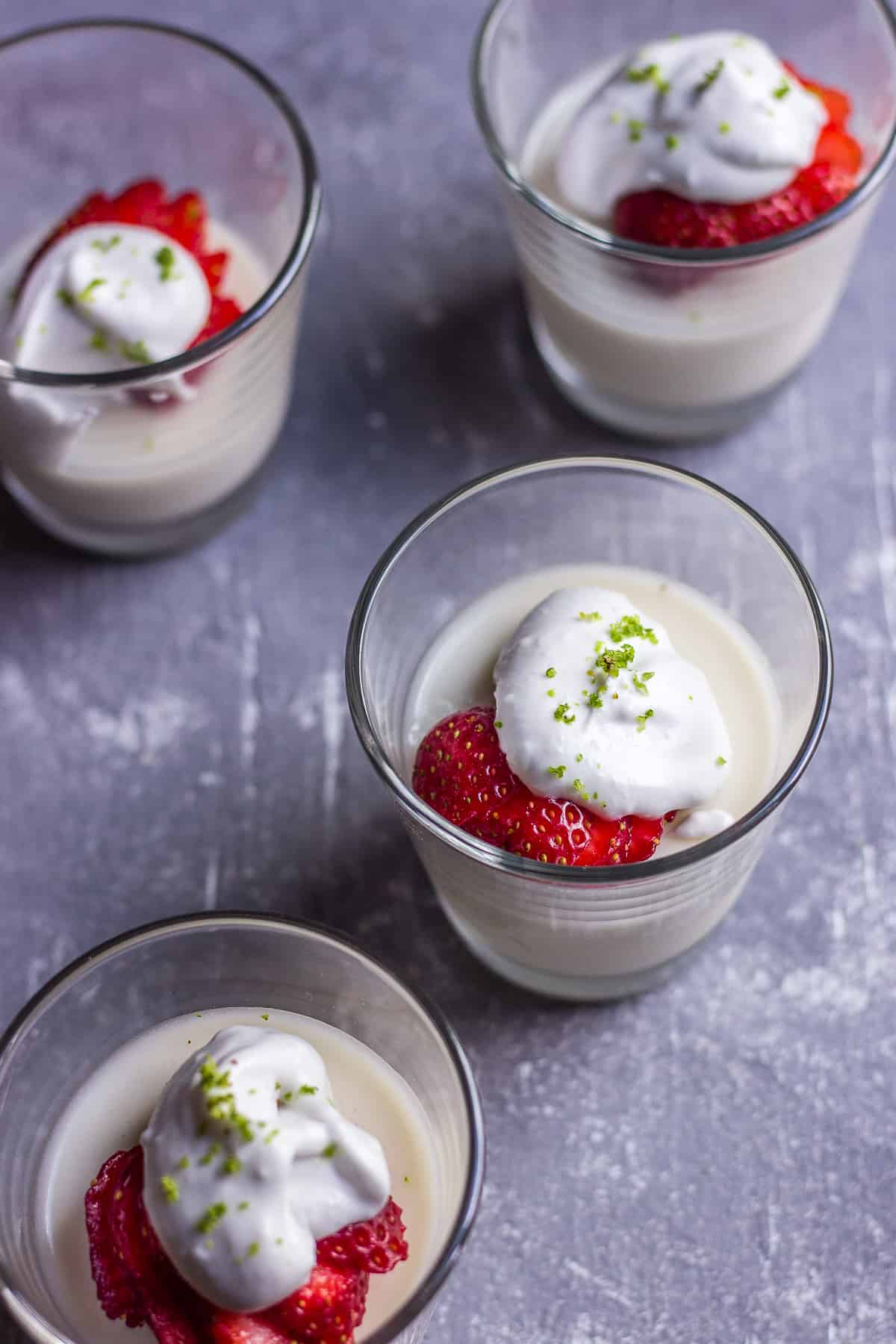 Four dessert cups with a white pudding with strawberries and cream on top with a green garnish.