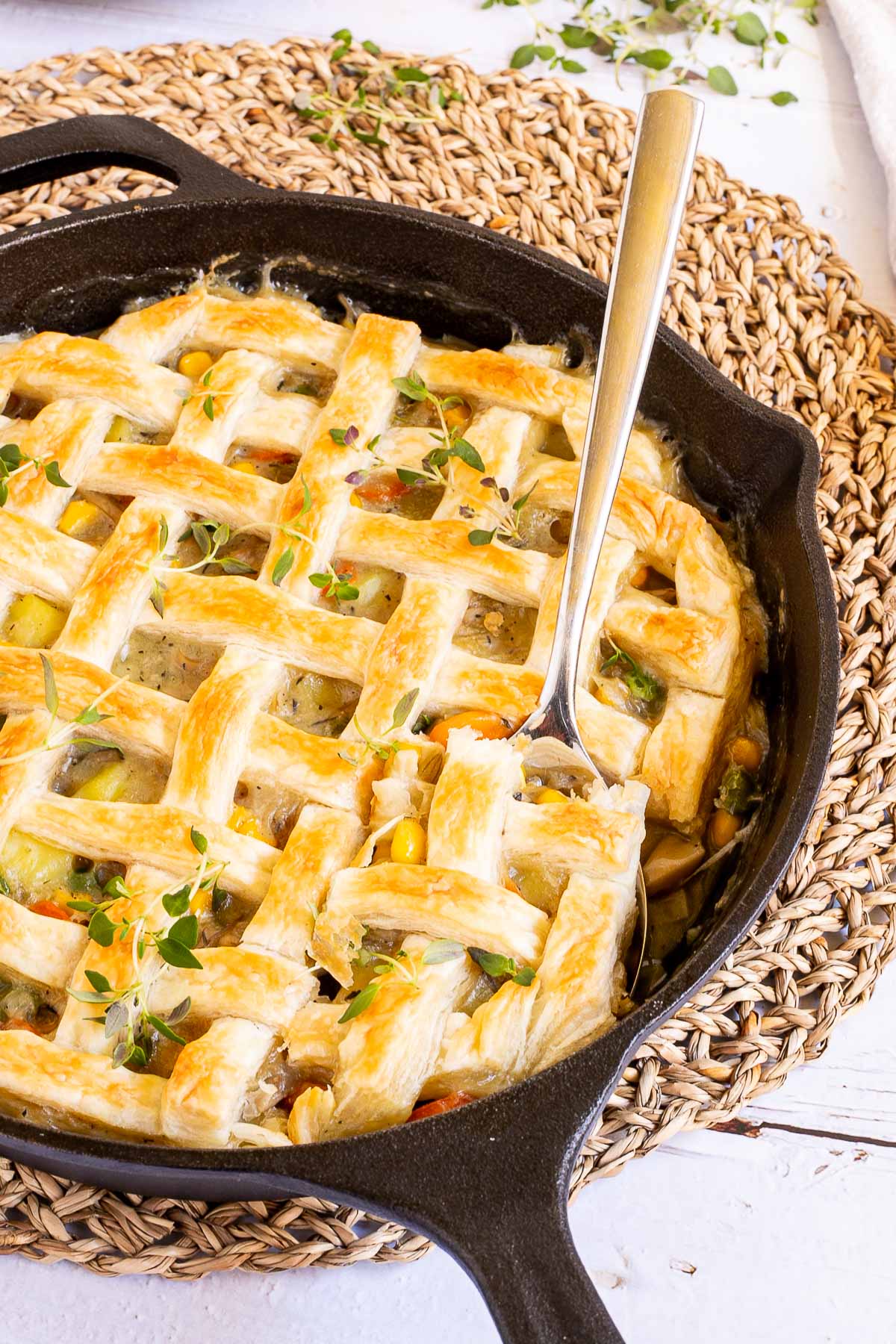 Cast iron skillet with veggie stew topped with puff pastry in a lattice pattern. A large serving spoon is placed in the middle.