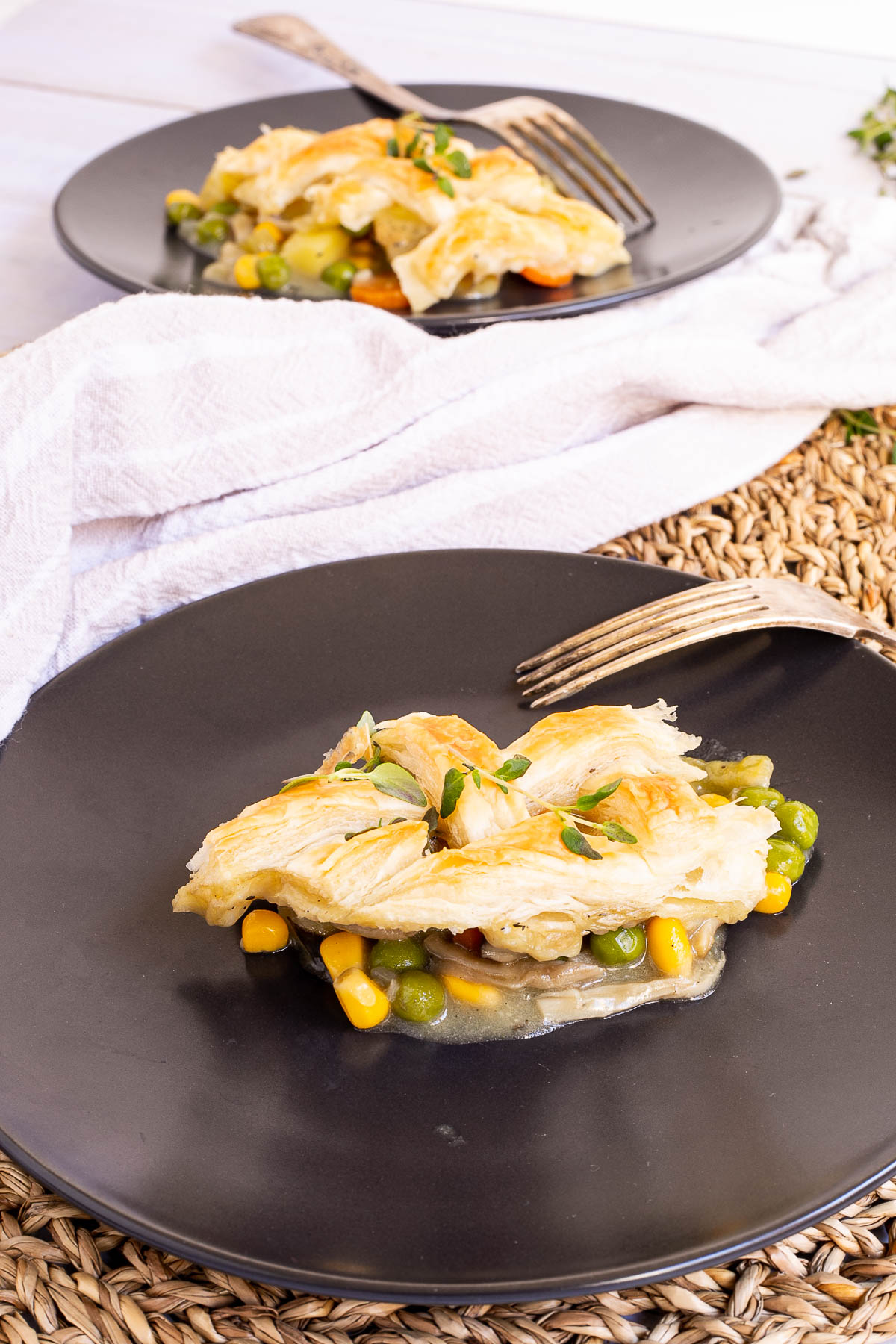 A serving of pot pie is placed on black plate. Another serving is right behind it. The part of the lattice pattern and some veggies are visible.