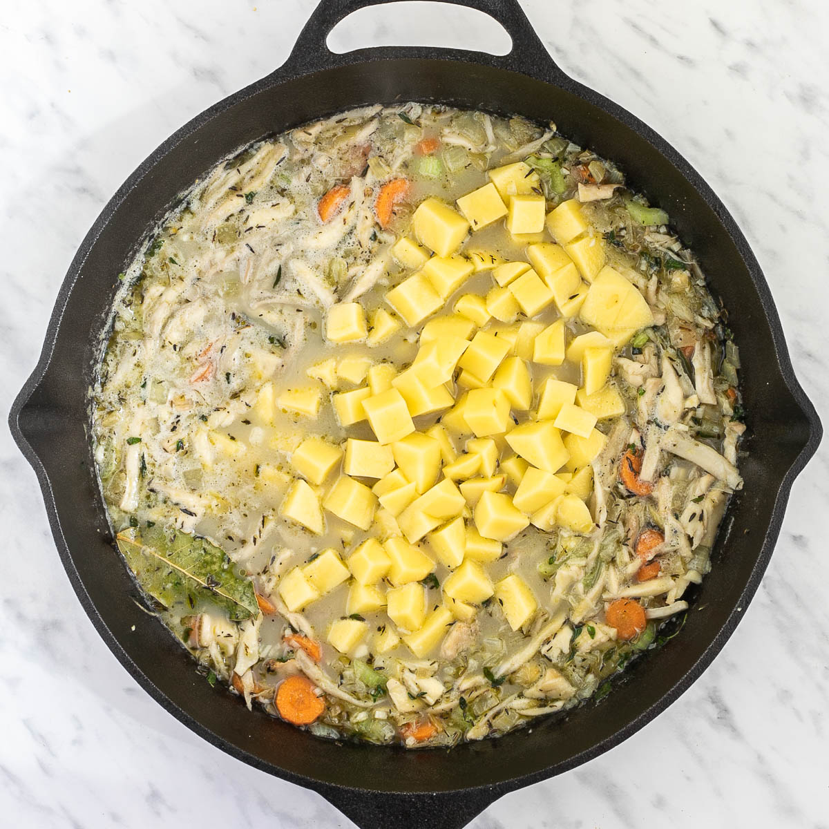 Cast iron skillet with light broth-like liquid with sliced carrots, chopped onion, garlic, celery, shredded king oyster mushrooms, bay leaves, thyme and a heap of potatoes in the middle.