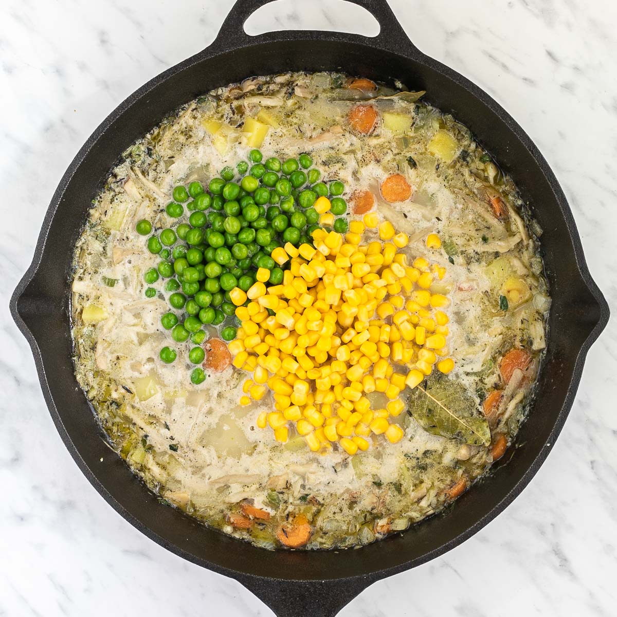 Cast iron skillet with light broth-like liquid with sliced carrots, chopped onion, garlic, celery, potatoes, shredded king oyster mushrooms, bay leaves, thyme and a heap of corn and green peas in the middle.