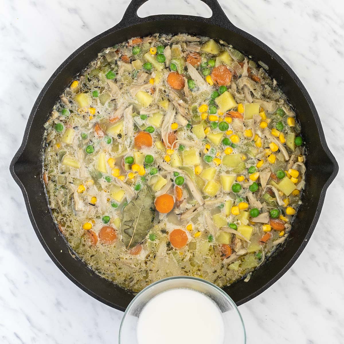 Cast iron skillet with light broth-like liquid with sliced carrots, chopped onion, garlic, celery, potatoes, corn, peas, shredded king oyster mushrooms, bay leaves, thyme. A small bowl of white slurry is placed next to it.